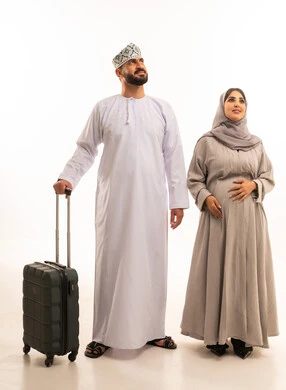 Looking up at something, having fun during the vacation, enjoying travel and summer trips, a portrait of an Arab Gulf Omani man wearing a dishdasha and a kumma holding a travel bag with his wife, the concept of pregnancy and childbirth, a full-body image, white background.