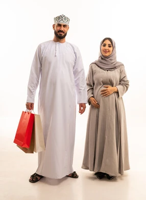 التبضع وأخذ المستلزمات الضرورية،بورترية لرجل عربي خليجي عماني يرتدي الدشداشة و الكمة  يحمل أكياس ورقية ملونه برفقة زوجته, استغلال العروض والتخفيضات الموسمية والجمعة البيضاء، مفهوم الحمل و الانجاب، التسوق من المولات التجارية، ايماءات السعاده و السرور، الجسم بالطول الكامل، خلفية بيضاء