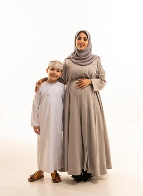 الأمومة والرعايه والاهتمام بالأبناء، مفهوم الحمل والانجاب، قضاء وقت ممتع مع الاطفال، بورترية لولد عربي خليجي عماني يرتدي الدشداشة و الكمه يقف برفقه والدته ينظران الى الكاميرا بتعابير السعادة و السرور، الجسم بالطول الكامل، خلفية بيضاء