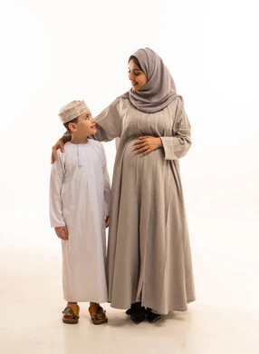 Happy family atmosphere filled with love and tenderness, spending enjoyable time with children, motherhood, care, and attention to children, the concept of pregnancy and childbirth, a portrait of an Arab Gulf Omani boy wearing a dishdasha and a kumma standing next to his mother, looking at each other with expressions of happiness and joy, full-body, white background.