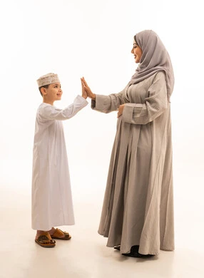 مفهوم الحب والألفة بين الابناء، إيماءات ضرب الكف، قضاء وقت ممتع باللعب مع الاولاد، بورترية لولد عربي خليجي عماني يرتدي الدشداشة و الكمة يشير بكف يده مقابل كف يد والدته، الجسم بالطول الكامل، خلفية بيضاء