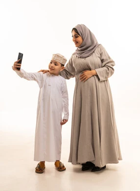 التقاط صور السيلفي لتوثيق اللحظات السعيدة، روابط المحبة والتماسك الأسري، بورترية لولد عربي خليجي عماني يرتدي الدشداشة و الكمه يقف برفقه والدته يمسك بيديه الهاتف المحمول،  مفهوم الحمل والانجاب، الجسم بالطول الكامل، خلفية بيضاء، سولوليت