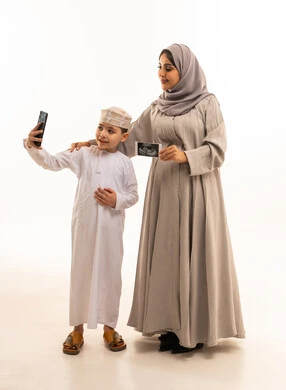 توثيق اللحظات السعيدة، روابط المحبة والتماسك الأسري، بورترية لولد عربي خليجي عماني يرتدي الدشداشة و الكمه يقف برفقه والدته يمسك بيديه الهاتف المحمول يقوم بالتقاط صوره سيلفي،  مفهوم الحمل والانجاب، امراه خليجيه محجبه تمسك بيديها كارت اشعه السونار، الجسم بالطول الكامل، خلفية بيضاء، سولوليت