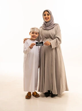 تربية الأبناء والاهتمام بهم، روابط المحبة والتماسك الأسري، بورترية لولد عربي خليجي عماني يرتدي الدشداشة و الكمه يقف برفقه والدته يحملون بايديهم كارت اشعه السونار، مفهوم الحمل والانجاب، تعابير السعادة و السرور، الجسم بالطول الكامل، خلفية بيضاء، سولوليت