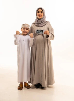 Achievement and goal realization, an Omani Gulf Arab boy wearing a dishdasha and kumma stands with his mother, raising his fists in the air with gestures indicating victory and triumph. An Omani Gulf Arab woman wearing a hijab and abaya holds ultrasound rays in her hands, representing the concept of pregnancy and childbirth, full-body image, white background, cellulite.