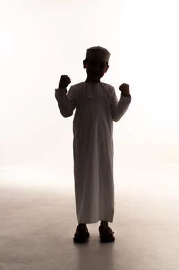 النظر الى الكاميرا بتعابير السعادة والحماس والسرور، مفهوم الانجاز وتحقيق الهدف،  صورة ظلية لولد عربي خليجي عماني مبتسم يرتدي الدشداشة والكمة يرفع قبضه يدية للاعلى بإيماءات الفوز والانتصار والنجاح, الجسم بالطول الكامل، خلفية بيضاء ، سولوليت