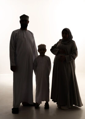 تعزيز المحبة والانسجام داخل الأسرة، رعاية الآباء بأبنائهم والاهتمام بهم، مفهوم الحمل والانجاب، بورتريه لرجل عربي خليجي عماني يرتدي الدشداشة و الكمة يقف برفقة زوجته و ابنه ينظران  للكاميرا بإيماءات السعادة، صورة الجسم بالطول الكامل، خلفية بيضاء، سولوليت
