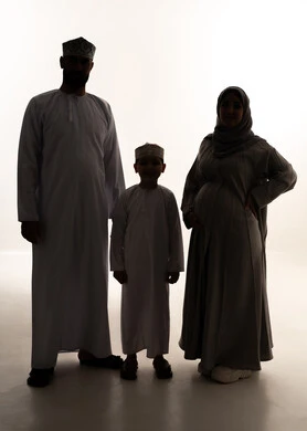 الترابط بين أفراد الأسرة واستقرار الحياة العائلية، رعاية الآباء بأبنائهم والاهتمام بهم، مفهوم الحمل والانجاب، بورتريه لرجل عربي خليجي عماني يرتدي الدشداشة و الكمة يقف برفقة زوجته و ابنه ينظران  للكاميرا بإيماءات السعادة، صورة الجسم بالطول الكامل، خلفية بيضاء، سولوليت