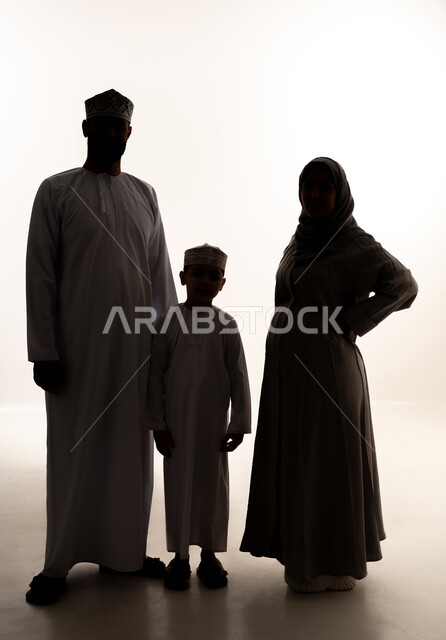 الترابط الأسري والحفاظ على الاستقرار العائلي، رعاية الآباء بأبنائهم والاهتمام بهم، مفهوم الحمل والانجاب، بورتريه لرجل عربي خليجي عماني يرتدي الدشداشة و الكمة يقف برفقة زوجته و ابنه ينظران  للكاميرا بإيماءات السعادة، صورة الجسم بالطول الكامل، خلفية بيضاء، سولوليت
