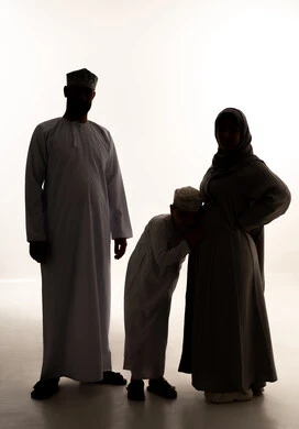 مفهوم انتظار قدوم المولود، مفهوم الحمل والانجاب، النظر إلى الكاميرا بإيماءات الحماس، صورة ظلية لولد عربي خليجي عماني يرتدي الدشداشة و الكمة يضع اذنه على بطن والدته برفقة ابيه، الجسم بالطول الكامل، خلفية بيضاء، سولوليت