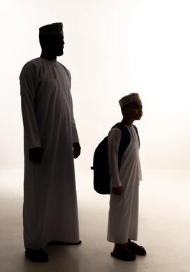 Caring for and attending to children, preparing to start a new school year, the back-to-school season in Oman, the feeling of safety and love with parents, a side silhouette of an Omani Gulf Arab boy wearing a dishdasha and kumma carrying a backpack alongside his father, full body, white background, silhouette.