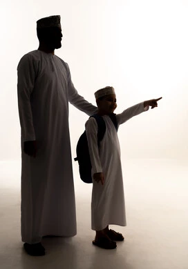 الاشارة الي شيء ما ، الاستعداد للعام الدراسي، مفهوم الاعتناء بالأبناء والاهتمام بهم، موسم العودة للمدارس في عمان، الشعور بالأمان والحب مع الآباء، صورة ظليةلولد عربي خليجي عماني يرتدي الدشداشة و الكمة يحمل حقيبة الظهر برفقة والده، الجسم بالطول الكامل، خلفية بيضاء ، سولوليت