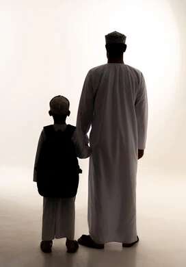 Preparation for the school year, the concept of caring for and paying attention to children, the back-to-school season in Oman, the feeling of safety and love with parents, a silhouette from behind of an Arab Gulf Omani boy wearing a dishdasha and kumma carrying a backpack accompanied by his father, full-body, white background, silhouette.