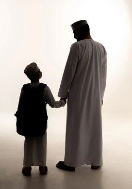 مفهوم الاعتناء بالأبناء والاهتمام بهم، موسم العودة للمدارس في عمان، الشعور بالأمان والحب مع الآباء، صورة ظلية من الخلف لولدعربي خليجي عماني يرتدي الدشداشة و الكمة يحمل حقيبة الظهر برفقة والده، الاستعداد للعام الدراسي، الجسم بالطول الكامل، خلفية بيضاء ، سولوليت
