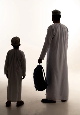 موسم العودة للمدارس في عمان، الشعور بالأمان والحب مع الآباء، مفهوم الاعتناء بالأبناء والاهتمام بهم، صورة ظلية من الخلف لرجل عربي خليجي عماني يرتدي الدشداشة و الكمة يمسك حقيبة الظهر برفقة ابنه، الاستعداد للعام الدراسي، الجسم بالطول الكامل، خلفية بيضاء ، سولوليت