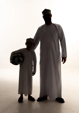 Sharing fun moments with children, the concept of fitness and physical agility, enjoying engaging in youth activities and sports, a silhouette of an Omani Gulf Arab boy wearing a dishdasha and kumma holding a football in his hands alongside his father, spending enjoyable times, full-body, white background, silhouette.