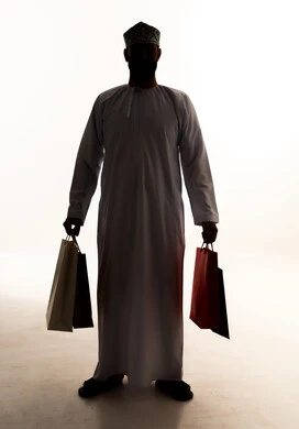 متعة التسوق في المولات التجارية، الاستفادة من العروضات والتخفيضات، شراء الحاجات والهدايا، النظر إلى الكاميرا بإيماءات البهجة والسرور، صورة ظلية لرجل عربي خليجي عماني يرتدي الدشداشة و الكمة يحمل بيديه اكياس ورقية، خلفية بيضاء ، سولوليت