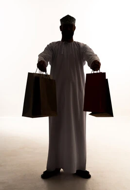 Taking advantage of offers and discounts, buying necessities and gifts, the joy of shopping in commercial malls, looking at the camera with gestures of joy and happiness, a silhouette of an Arab Gulf Omani man wearing a dishdasha and kumma holding paper bags in his hands, white background, silhouette.