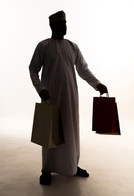 شراء الحاجات والهدايا، الاستفادة من العروضات والتخفيضات، متعة التسوق في المولات التجارية، صورة ظلية  لرجل عربي خليجي عماني يرتدي الدشداشة و الكمة يحمل بيديه اكياس ورقية، خلفية بيضاء ، سولوليت