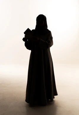 بيع وشراء العقارات، السعي لامتلاك بيت جديد، الروابط الأسرية القوية،صورة ظلية لامراه عربيه خليجيه سعودية ترتدي العباءة و الحجاب تحمل بيديها مجسم خشبي صغير لمنزل الجسم بالطول الكامل، خلفية بيضاء ، سولوليت