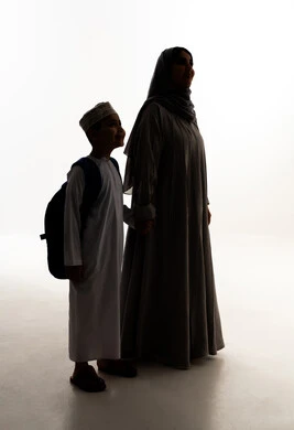 النظر الى شيء ما، الشعور بالأمان والحب مع الامهات ، موسم العودة للمدارس في سلطنه عمان، مفهوم الاعتناء بالأبناء والاهتمام بهم، صورة ظلية لولد عربي خليجي عماني يرتدي الدشداشة و الكمة و الحقيبة المدرسية يقف برفقة والدته، الاستعداد للعام الدراسي، الجسم بالطول الكامل، خلفية بيضاء ، سولوليت