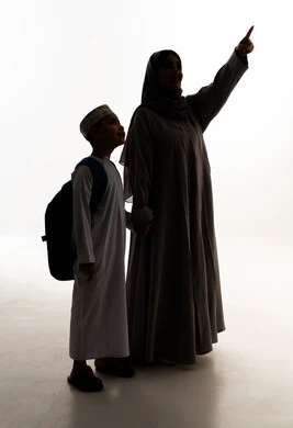 The gesture and looking at something, the feeling of safety and love with mothers, the back-to-school season in the Sultanate of Oman, the concept of caring for and paying attention to children, a silhouette of an Arab Gulf Omani boy wearing a dishdasha and kumma and carrying a school bag standing with his mother, preparing for the school year, full-body, white background, silhouette.
