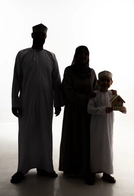 السعي لامتلاك بيت جديد، بيع وشراء العقارات، صورة ظلية لولد عربي خليجي عماني يرتدي الدشداشة و الكمة يحمل بيديه مجسم خشبي صغير لمنزل برفقة والدية، إيماءات السرور والسعادة، خلفية بيضاء ، سولوليت