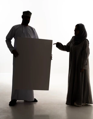 الاشارة الى اللوحة، استخدام لوحات توضيحية، مفهوم الإعلان والتسويق لمكان ما، الترويج للخدمات والمنتجات، موك اب مربع صغير، صورة ظلية لزوجان عربيان خليجيان عمانيان يرتديان الزي التقليدي يمسكان بيديهم لوحة بيضاء فارغة، لوح إعلاني فارغ ،الجسم بالطول الكامل، خلفية بيضاء، سولوليت