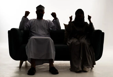 صورة ظلية لزوجان عربيان خليجيان عمانيان يجلسان على الاريكة يقومان برفع  يديهم مضمومه الكفين يقومان بالتشجيع, إيماءات وجه ويدين تدل على السعاده, التفاعل والإندماج, مفهوم الفوز والإنتصار, أنديه وتشجيع, خلفية بيضاء، سولوليت