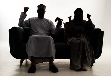 Spending enjoyable times, a silhouette of an Arab Gulf Omani man wearing a dishdasha and kumma sitting on the couch with his wife holding a game controller, playing video games, various facial and hand gestures indicating victory and triumph, white background, silhouette.