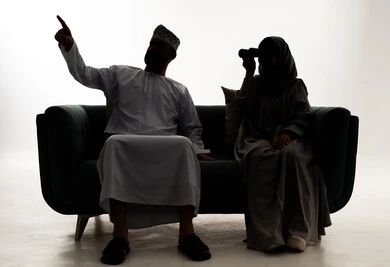التخطيط المستقبلي للأهداف و الطموحات، الاشارة الى شيء ما، حب الاستكشاف والمغامرة، رؤية الاشياء البعيدة عبر التلسكوب، صورة ظلية لامرأة عربية خليجية عمانية محجبة ترتدي العباءة الملونة تجلس على الاريكة برفقة زوجها تنظر من خلال منظار ثنائي العينين, خلفية بيضاء ، سولوليت