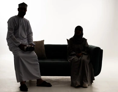 تقنيات حديثة الكترونية وأجهزة تكنولوجية،المتعة والتسلية بالمشاهدة عبر نظارة الواقع الافتراضي، صورة ظلية لرجل عربي خليجي عماني يرتدي الدشداشة و الكمة يجلس على الاريكة برفقة زوجته يمسك بيديه نظارات الفي ار، النظر الى الكاميرا بايماءات السعادة و السرور، خلفية بيضاء ، سولوليت