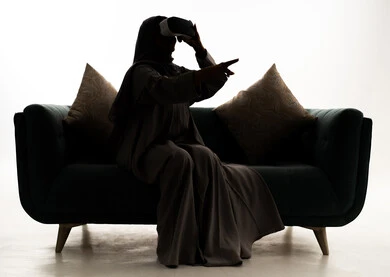 Integration and simulation of the imaginary world, referring to something, using modern electronic technology, a silhouette of a Saudi Gulf Arab woman wearing a hijab and an abaya sitting on a sofa holding 3D glasses (VR) in her hand, enjoyment and entertainment through viewing via virtual reality glasses, white background, silhouette.