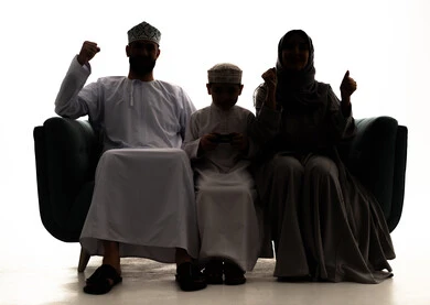 تحقيق النجاح والانتصار في المنافسات، صورة ظلية لعائلة عربيه خليجيه عمانية ترتدي الزي التقليدي يجلسون على الاريكه يقومون برفع ايديهم للاعلي بايماءات السرور والسعاده، ولد عماني يقوم بامساك عصا التحكم، مفهوم الفوز بالبطولات، ألعاب البلايستيشن الترفيهية والممتعة ، خلفية بيضاء ، سولوليت