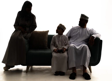 مفهوم الحب و الترابط الاسري، صورة ظلية لعائلة عربية خليجيه عمانية ترتدي الزي التقليدي يجلسون على الاريكة ينظرون الى بعض بايماءات السعادة و السرور، ولد عماني يقوم بامساك عصا التحكم،  قضاء أوقات عائلية ممتعة، خلفية بيضاء ، سولوليت