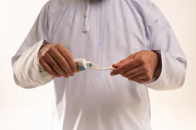 Using a toothbrush and toothpaste, the concept of healthy habits, maintaining and continuously cleaning teeth, a bright white smile, daily oral hygiene routine, avoiding cavities and gum diseases, a close-up portrait of an Arab Omani Gulf man wearing a dishdasha and kumma brushing his teeth, white background.
