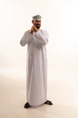 المحافظة على الأناقة والنظافة الشخصية، الاهتمام بالمظهر الخارجي، بورتريه لرجل عربي خليجي عماني يرتدي الدشداشة والكمة يقوم بإزالة شعر الوجه باستخدام شفرة الحلاقة الرجالية ،صورة الجسم بالطول الكامل، خلفية بيضاء