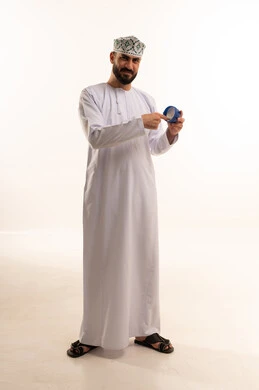 Looking at the camera with expressions of joy, the concept of caring for the freshness and improvement of the skin, moisturizing, cleansing, and brightening the skin, skincare routine, using youthful cosmetic ingredients, a portrait of an Arab Gulf Omani man wearing a dishdasha and kumma applying moisturizer, full-body shot, white background.