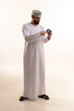 مفهوم الإهتمام بنضارة وتحسين البشرة، ترطيب وتنظيف وتفتيح الجلد، روتين العناية بالشرة، استخدام مكونات تجميلية شبابية، بورتريه لرجل عربي خليجي عماني يرتدي الدشداشة والكمة يضع كريم المرطب ، الجسم بالطول الكامل، خلفية بيضاء