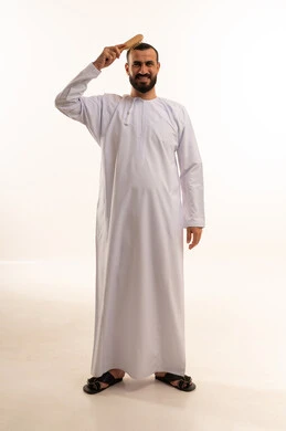 تعابير وإيماءات تدل على السعادة والسرور، مفهوم العناية والاهتمام بالجمال والأناقة والمظهر الخارجي، بورتريه دلرجل عربي خليجي سعودي يرتدي الثوب الابيض يقوم بتصفيف شعره باستخدام فرشاة الشعر ( المشط )، الجسم بالطول الكامل، خلفية بيضاء