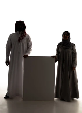 استخدام لوحات توضيحية، مفهوم الإعلان والتسويق لمكان ما، الترويج للخدمات والمنتجات، موك اب مربع صغير، صورة ظلية لزوجان عربيان خليجيان سعوديان يرتديان الزي التقليدي  يمسكان بيديهم لوحة بيضاء فارغة، لوح إعلاني فارغ ،الجسم بالطول الكامل، خلفية بيضاء، سولوليت