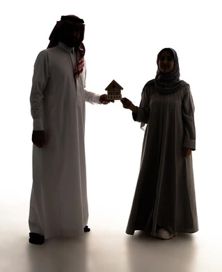 السعي لامتلاك بيت جديد، بيع وشراء العقارات،  صورة ظلية لرجل عربي خليجي سعودي يرتدي الثوب الابيض و الشماغ يحمل بيديه مجسم خشبي صغير لمنزل برفقة زوجته، إيماءات السرور والسعادة، سولوليت