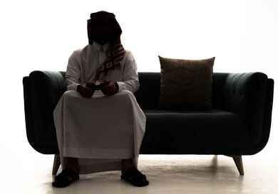 Modern electronic entertainment technologies, interaction and immersion with PlayStation games, a silhouette of an Arab Saudi Gulf man wearing a ghutrah and traditional thobe sitting on the couch holding a game controller playing video games, white background, silhouette.