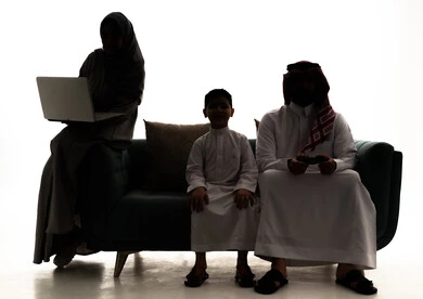 The use of modern technological devices, entertaining and enjoyable PlayStation games, a silhouette of a Saudi Arabian Gulf man wearing a white thobe and a ghutrah sitting with his son and wife holding a game controller in his hands, a veiled Saudi woman using a laptop, white background, silhouette.