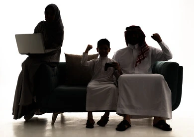 مفهوم الفوز بالبطولات، تحقيق النجاح والانتصار في المنافسات، صورة ظلية لاب عربي خليجي سعودي يرتدي الثوب الابيض و الشماغ يجلس على الاريكة  يقوم برفع يدية  للاعلي بايماءات السرور والسعاده برفقة ابنه، امراة سعودية ترتدي العباءة و الحجاب تستخدم الحاسب المحمول، خلفية بيضاء ، سولوليت