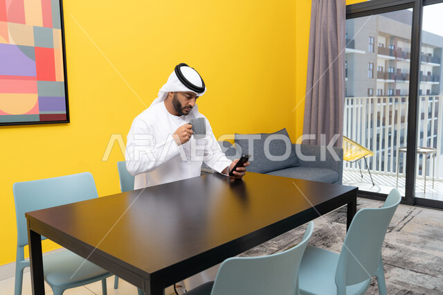 تبادل الأحاديث والتواصل مع الأهل والأصدقاء، رجل عربي خليجي اماراتي يرتدي الكندورة والغترة البيضاء يجلس خلف الطاولة ويمسك بيده الهاتف المحمول، استخدام جهاز تقني حديث ومتطور، العمل عن بعد، تصفح التطبيقات وبرامج التواصل الاجتماعي، الاستمتاع بشرب القهوة في المنزل