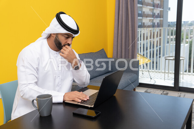 استخدام جهاز تقني حديث ومتطور، رجل عربي خليجي اماراتي يرتدي الكندورة والغترة البيضاء يجلس خلف الطاولة يشرب القهوة ويستخدم الحاسوب المحمول، إنجاز مهام العمل عن بعد، إدارة شؤون الأعمال وتنظيمها عبر اللابتوب، الاستمتاع بالمشروبات الساخنة في المنزل