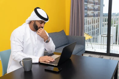 استخدام جهاز تقني حديث ومتطور، رجل عربي خليجي اماراتي يرتدي الكندورة والغترة البيضاء يجلس خلف الطاولة يشرب القهوة ويستخدم الحاسوب المحمول، إنجاز مهام العمل عن بعد، إدارة شؤون الأعمال وتنظيمها عبر اللابتوب، الاستمتاع بالمشروبات الساخنة في المنزل