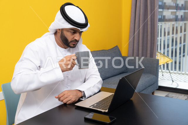 إدارة شؤون الأعمال وتنظيمها عبر اللابتوب، الاستمتاع بالمشروبات الساخنة في المنزل، استخدام جهاز تقني حديث ومتطور، رجل عربي خليجي اماراتي يرتدي الكندورة والغترة البيضاء يجلس خلف الطاولة يشرب القهوة ويستخدم الحاسوب المحمول، إنجاز مهام العمل عن بعد