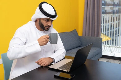 إدارة شؤون الأعمال وتنظيمها عبر اللابتوب، الاستمتاع بالمشروبات الساخنة في المنزل، استخدام جهاز تقني حديث ومتطور، رجل عربي خليجي اماراتي يرتدي الكندورة والغترة البيضاء يجلس خلف الطاولة يشرب القهوة ويستخدم الحاسوب المحمول، إنجاز مهام العمل عن بعد